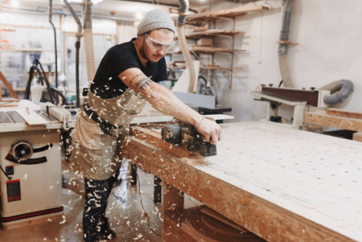 A carpenter wearing safety goggles and a beanie uses an electric planer on a wooden table, producing flying wood shavings in a workshop setting.